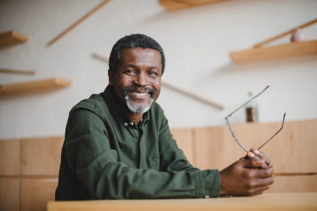 A smiling older Black man with a beard sits at a table, holding a pair of glasses.