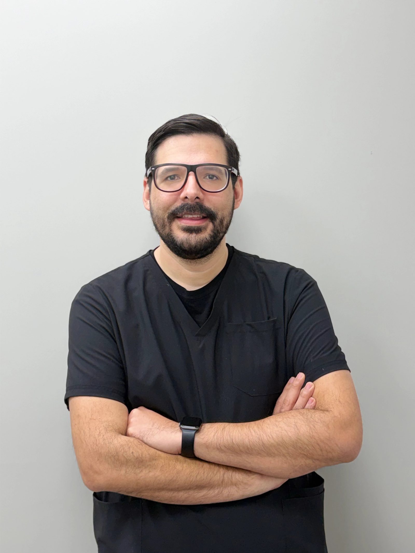 Man in black scrubs and glasses, arms crossed, smiling.