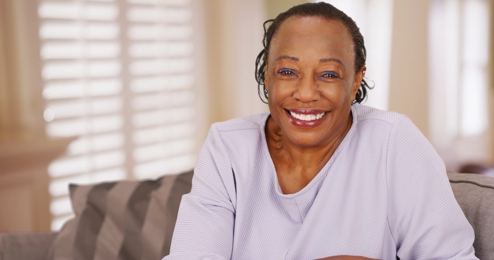 A smiling older Black woman in a light purple shirt sits indoors.