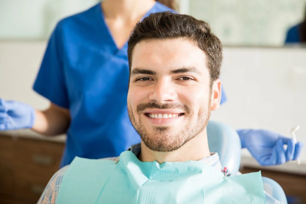 A smiling man in a dental chair with a dental bib, and a dental professional in the background.