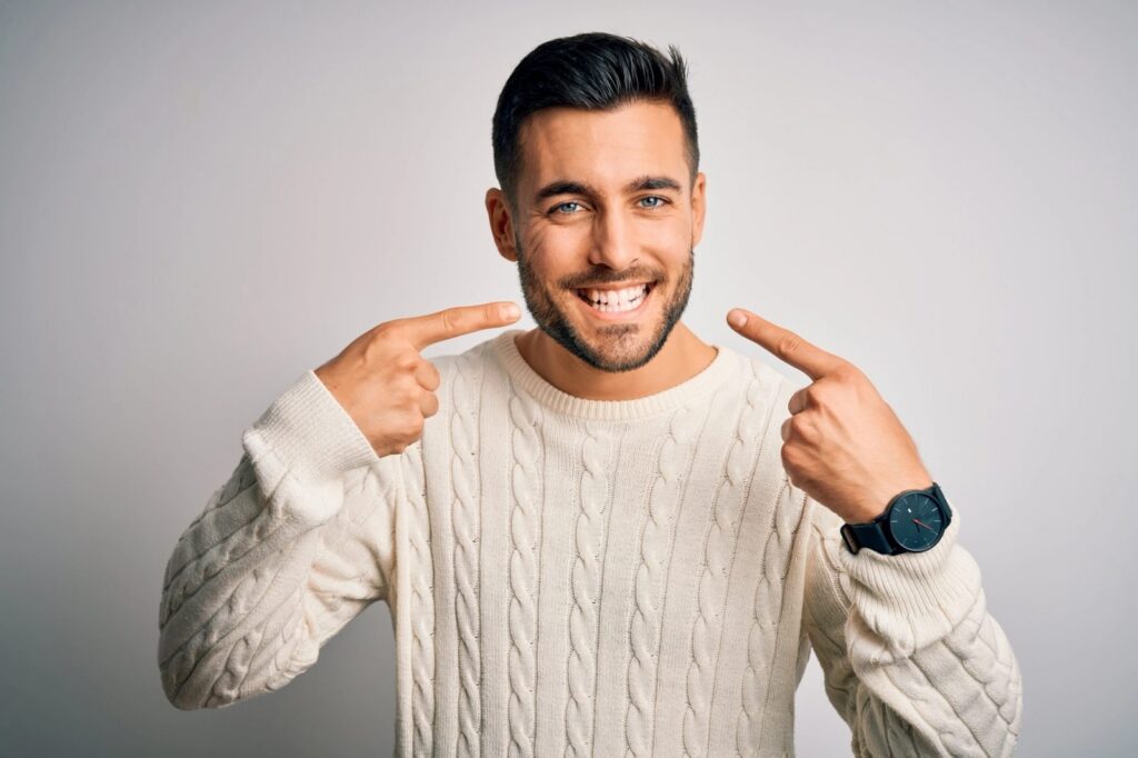 a young man wearing a white sweater is pointing at his teeth and smiling .